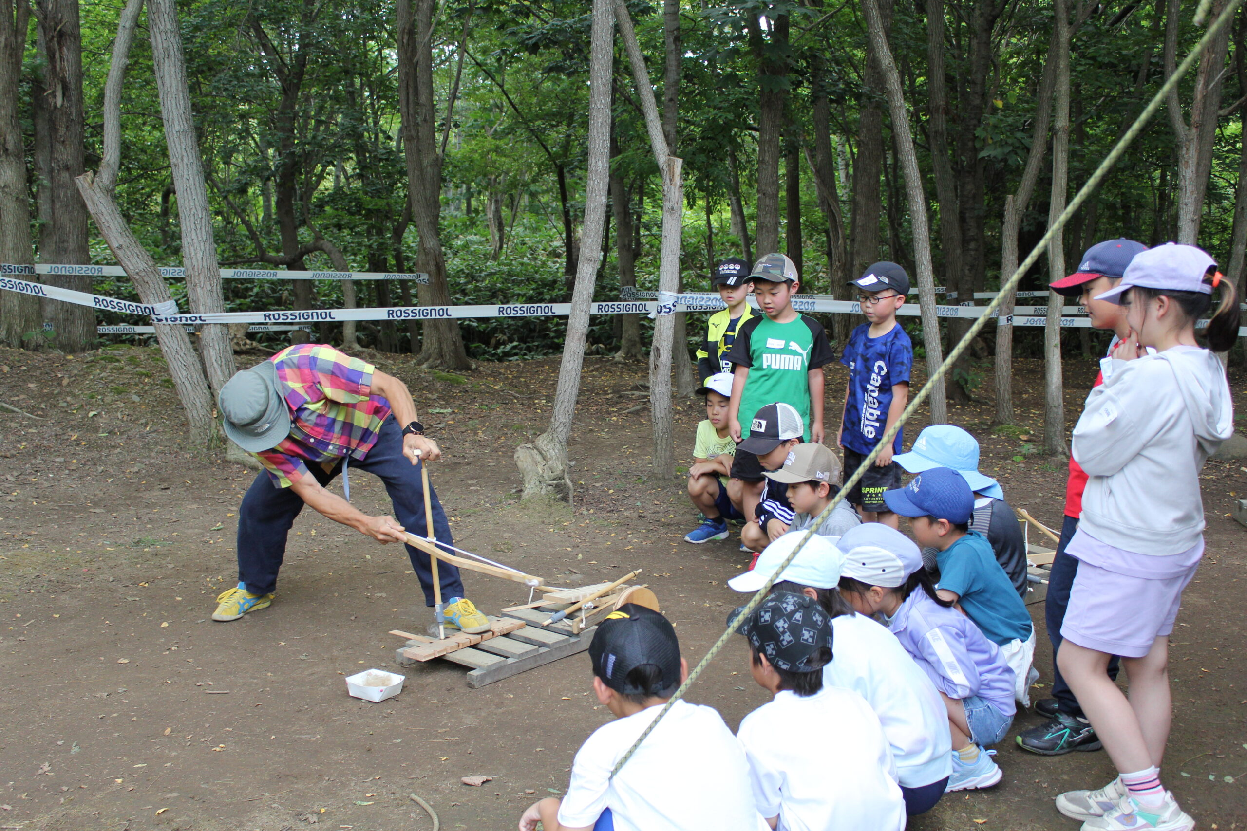 森の中で指導者の実演を子どもたちが見ながら自然体験活動に取り組んでいる様子